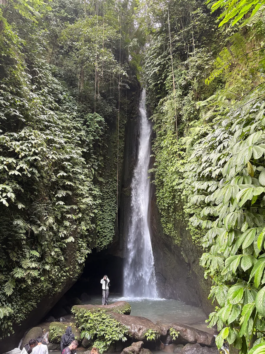 Leke Leke Waterfall in Tabanan, Bali, a tall thin waterfall dropping into a very narrow gorge with vine-draped walls, a single visitor standing at the base for scale.