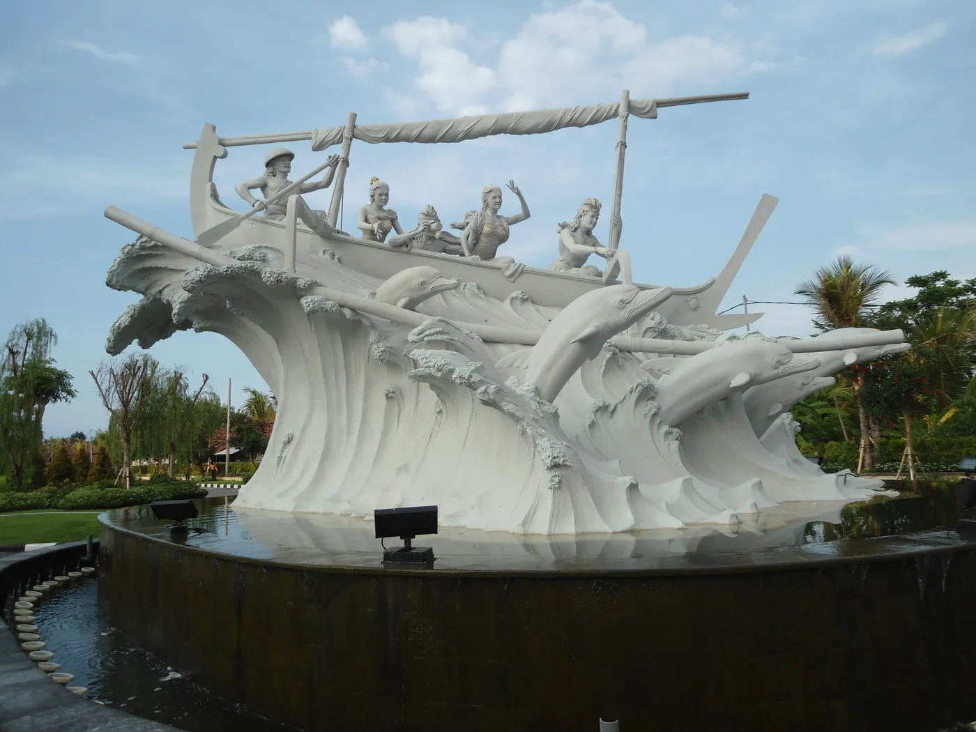 The dolphin monument at Temukus on Lovina beach in north Bali, a large white sculpture of a traditional outrigger boat with figures aboard riding a carved wave, dolphins leaping around the hull.