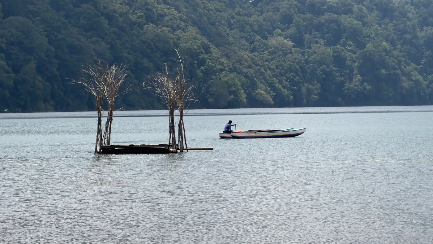 Lake Tamblingan in the old crater above Munduk, north Bali, a man paddling a wooden dugout canoe across still grey water beneath steep forested crater walls.
