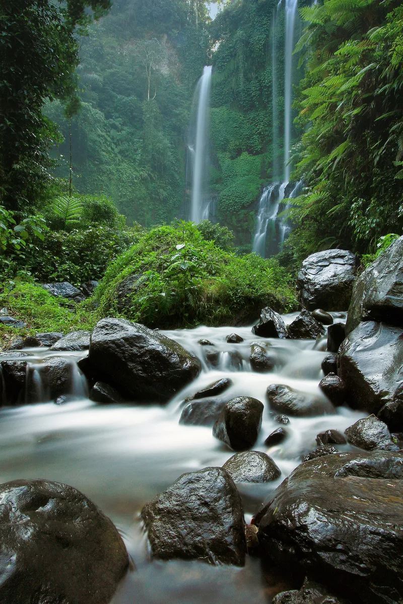Sekumpul Waterfall in Buleleng, north Bali, twin narrow falls dropping down a steep jungle cliff into a boulder-strewn river in the foreground.