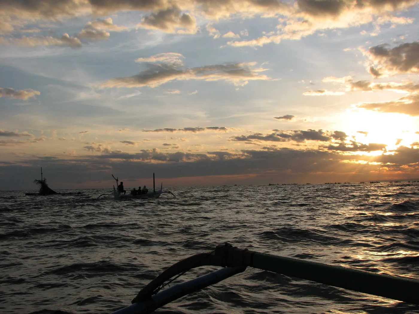 Sunrise seen from inside a jukung outrigger on a Lovina dolphin trip, the boat's bamboo outrigger arm in the foreground and a long line of other boats spread across the horizon.