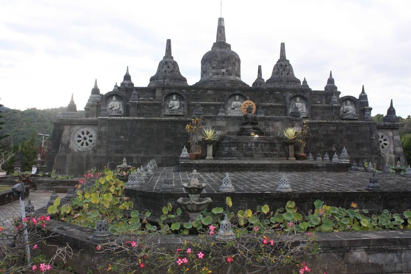 The miniature Borobudur at Brahmavihara-Arama Buddhist monastery near Banjar in north Bali, tiers of dark grey stone stupas with seated Buddha figures set into niches along the terrace wall.