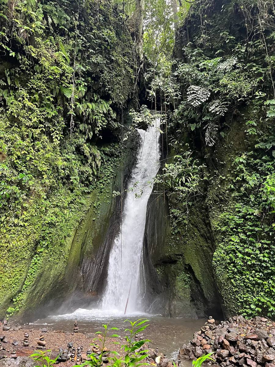 Belong Waterfall near Munduk, a single white column of water falling down a moss-covered rock gorge into a shallow brown pool with stacked stone cairns at its base.